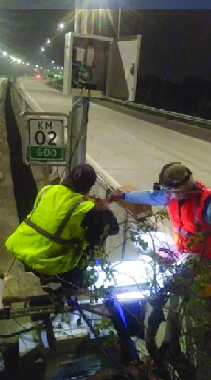 Dua teknisi bekerja di kaki pole cabinet perangkat di tepi jalan pada malam hari, dekat patok KM 02+600